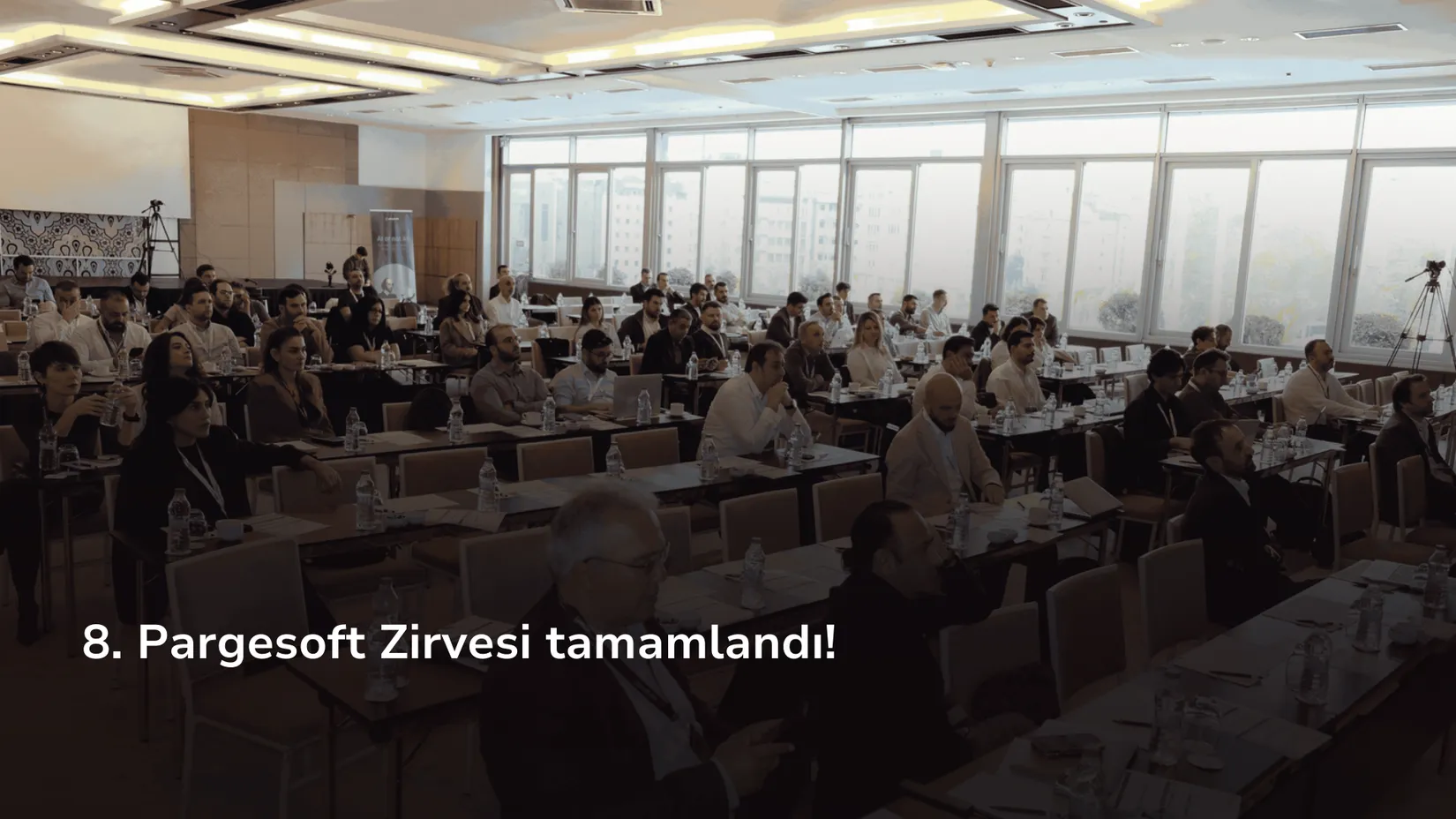 Bir konferans salonunda masalarda oturan çok sayıda katılımcı ekrandaki sunumu izliyor.