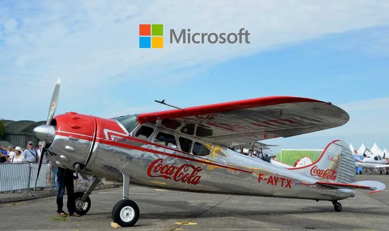 A vintage propeller plane in Coca-Cola livery parked on an airfield, with the Microsoft logo above it