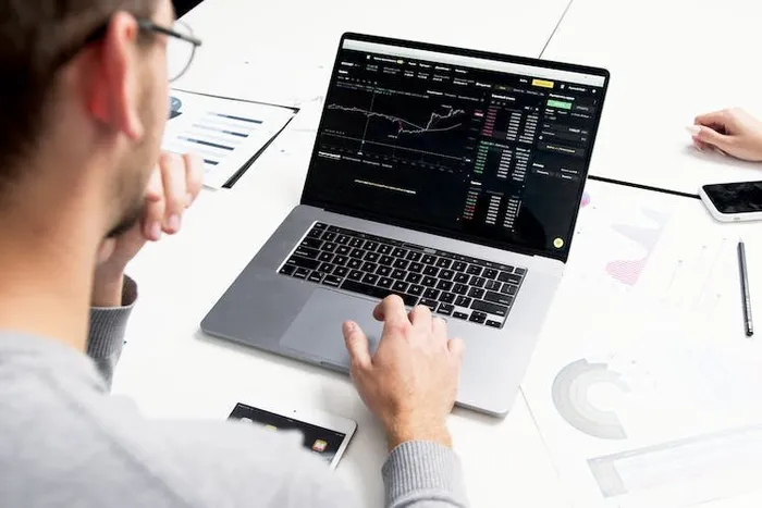 Laptop ekranında finansal grafik panosu; yanında basılı grafik ve raporlar, arkadan çekim.
