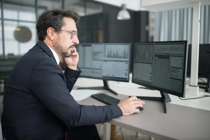 Takım elbiseli bir adamın telefonla konuşurken iki monitörde veri panosu incelemesi.