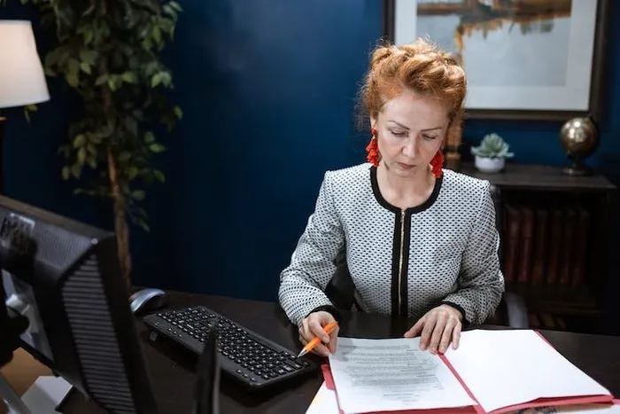 Koyu renkli bir ofis masasında kırmızı dosyadaki belgeyi imzalayan bir kadın.