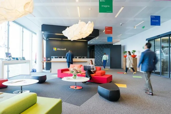 Duvarında Microsoft logosu bulunan bir ofis lobisi; kırmızı koltuklar ve renkli duyuru panoları.
