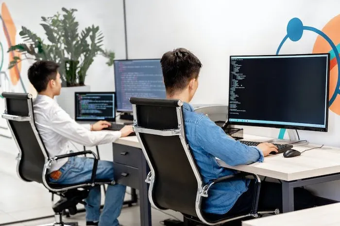 İki yazılımcı, kod görüntülenen çift monitörler başında sırtları dönük şekilde çalışıyor.