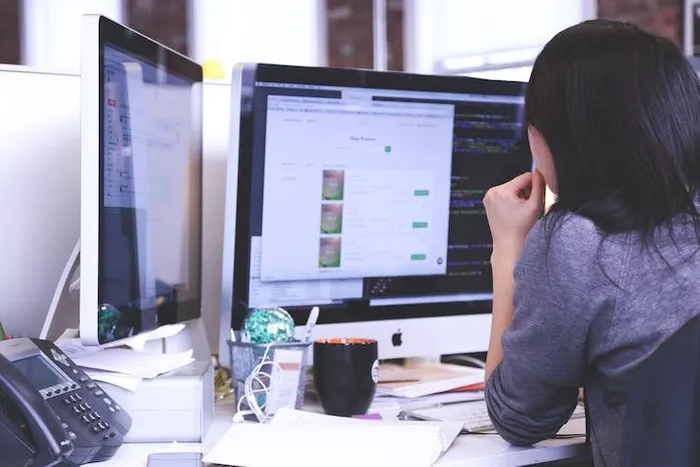 İki monitörlü masada, bir ekranda kod diğerinde ürün listesi görünen bir ofis çalışanı.