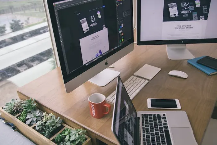 Ahşap masada iMac ve dizüstü bilgisayar ekranlarında web tasarım şablonları, yanında saksı bitkileri