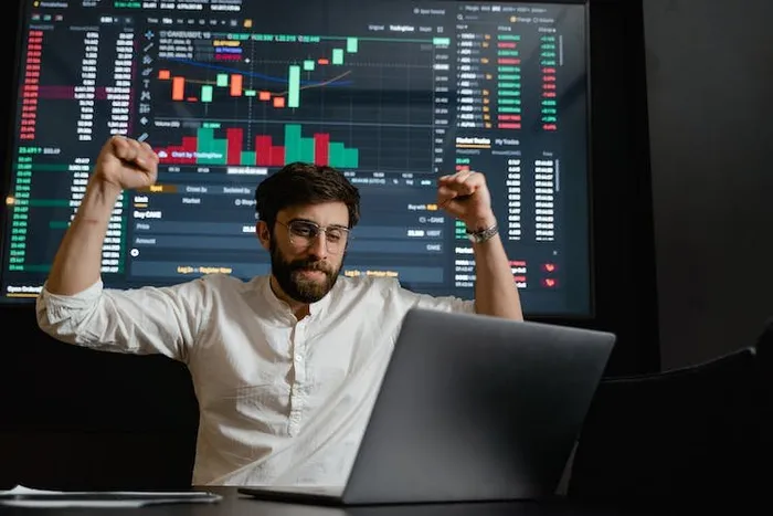 Bir adam laptop başında ellerini havaya kaldırıp sevinirken arkasındaki ekranda borsa grafiği var.