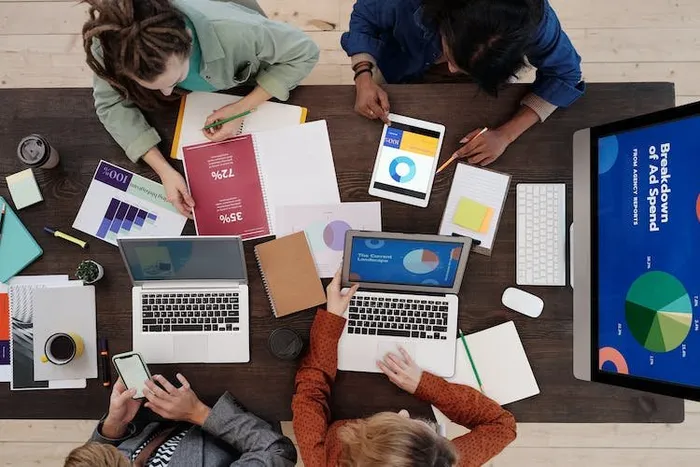 Masa başında dört kişi laptop ve tabletlerle çalışırken duvardaki ekranda reklam harcaması pasta grafiği var.