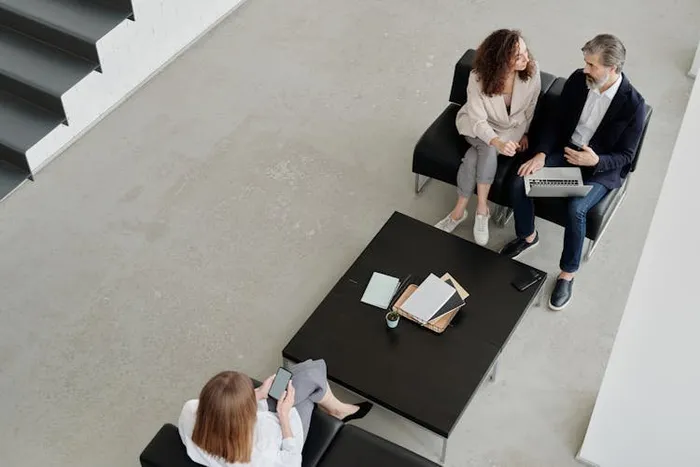 Üç kişi siyah koltuklarda oturmuş, önlerindeki masada defter ve telefonla sohbet ediyor.