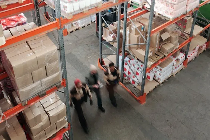 Kuşbakışı çekimde bir depoda üç çalışan, turuncu raflardaki koli yığınları arasında yürüyor