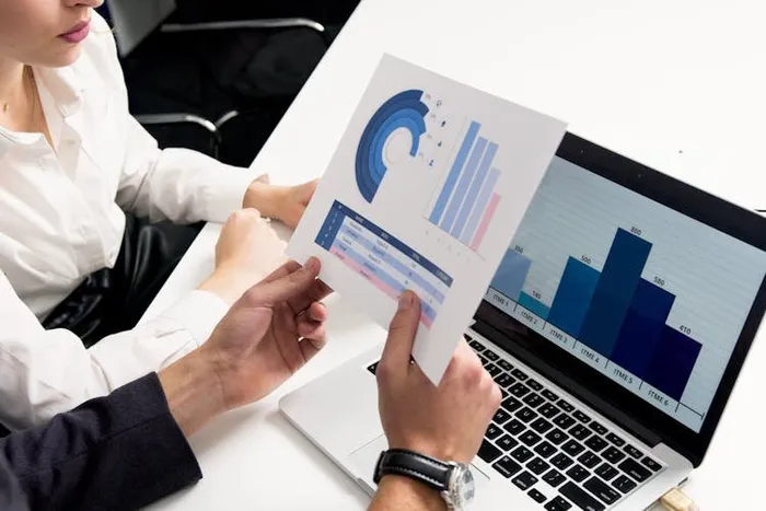 Bir kişi elindeki sütun grafikli raporu, ekranında aynı grafiği gösteren laptopun yanında tutuyor