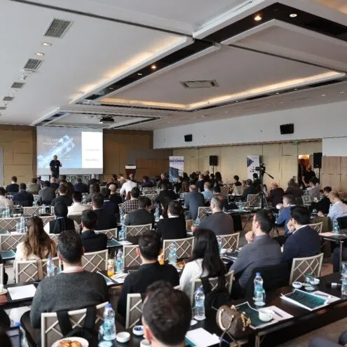 Masalarda oturan katılımcılarla dolu bir konferans salonu, önde sunum ekranı görülüyor