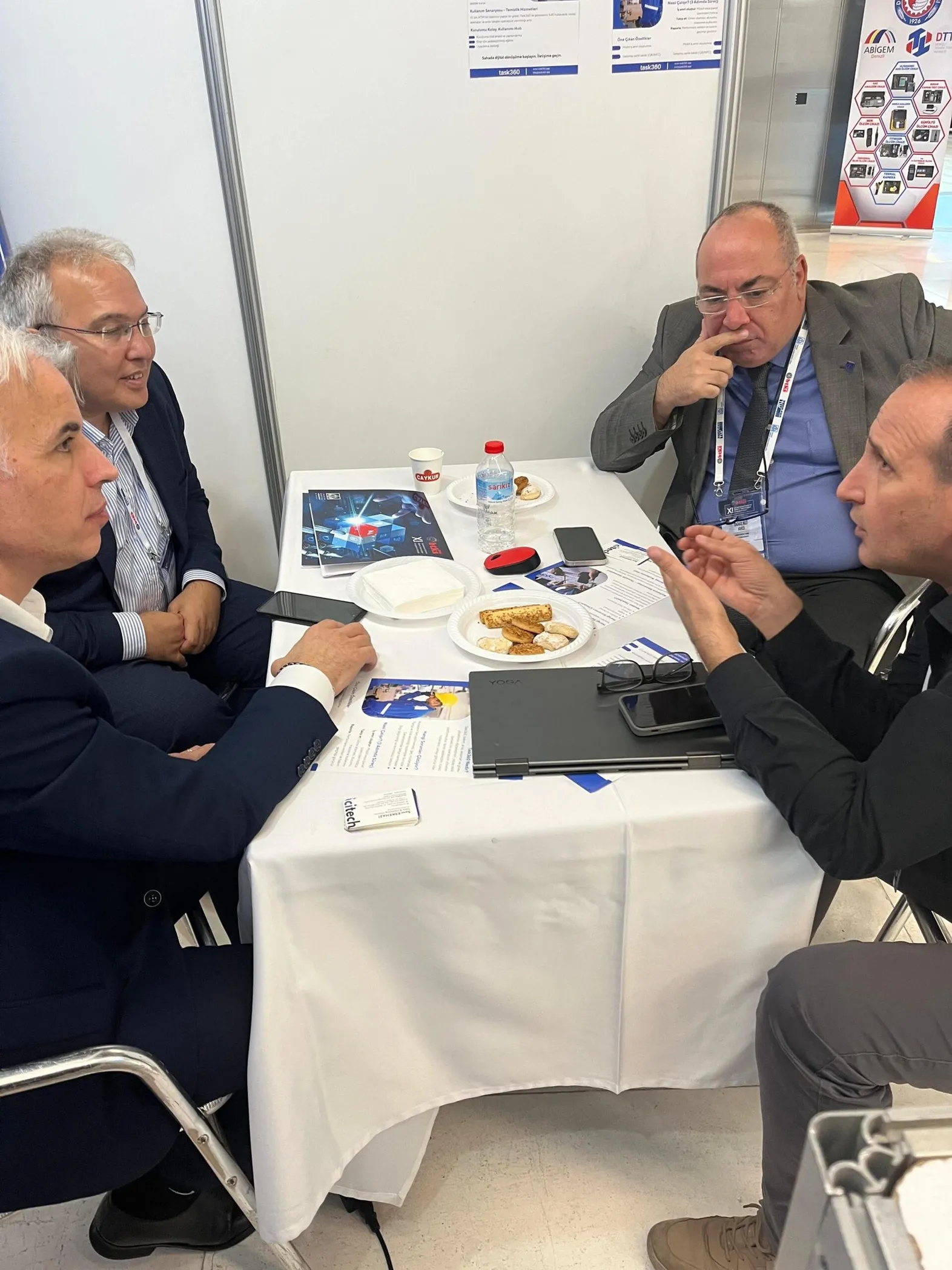 Task360 standında bir masada dört kişinin broşürler ve dizüstü bilgisayarla sohbet ettiği an
