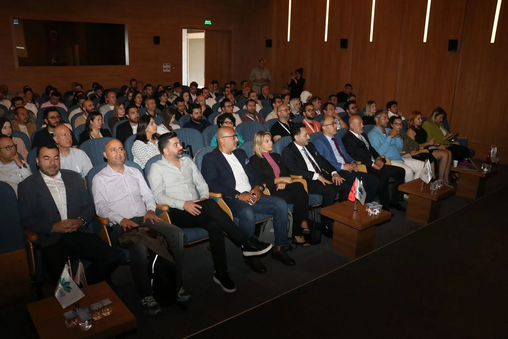Dijital Buluşmalar etkinliğinde mavi koltuklu geniş bir salonda oturan katılımcı kitlesi