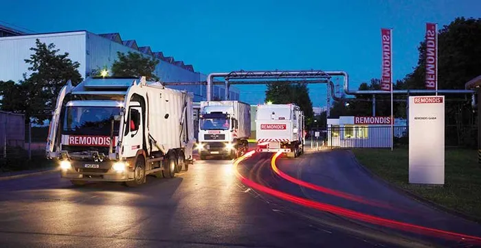 Alacakaranlıkta REMONDIS logolu çöp kamyonlarının bir tesis girişine yaklaşması